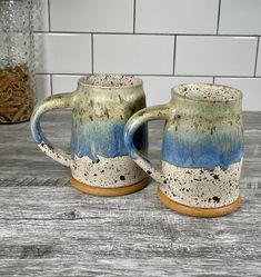 two coffee mugs sitting next to each other on top of a wooden table in front of a white tile wall