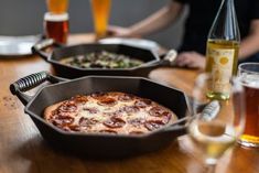 two pizzas in pans sitting on a table with beer and other food items