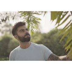 a man with a beard and tattoos standing in front of some trees looking up at the sky