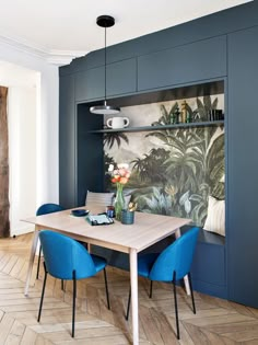 a dining room table with blue chairs next to a painting on the wall behind it