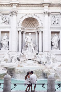 two people standing in front of a fountain with statues on the wall and behind them