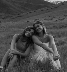 two women sitting in the grass hugging each other and smiling at the camera with mountains in the background