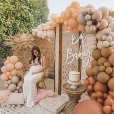 a pregnant woman sitting on a chair in front of a backdrop with balloons and cake