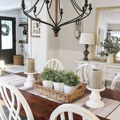 a chandelier hanging over a dining room table with white chairs and potted plants