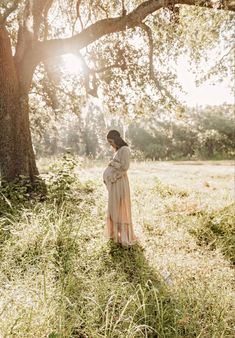 a woman standing under a large tree in the middle of a field with tall grass