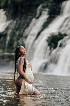 a pregnant woman sitting in the water next to a waterfall with her hands on her hips