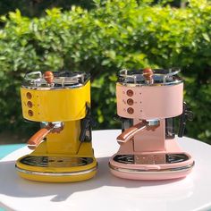 two colorful coffee machines sitting on top of a white table next to each other and green bushes in the background