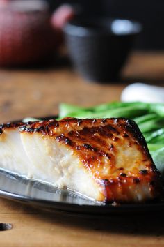 a piece of fish on a plate with asparagus and broccoli in the background