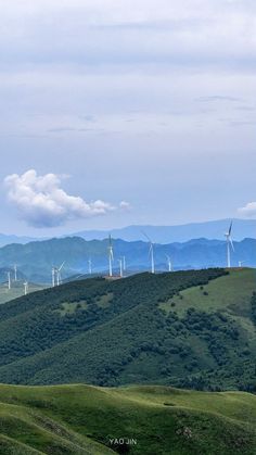 several wind mills on the side of a green hill with hills in the background and clouds in the sky