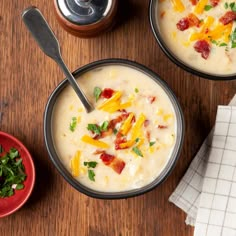 two bowls of soup with cheese and bacon in them on a wooden table next to a spoon