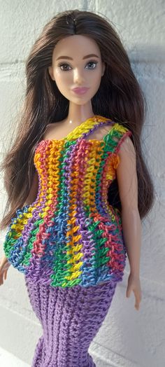 a doll with long hair wearing a colorful knitted top and skirt, standing next to a white brick wall