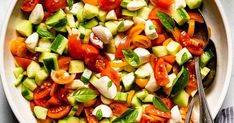 a white bowl filled with cucumber, tomatoes and other veggies next to a spoon