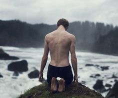 a shirtless man sitting on top of a cliff overlooking the ocean with his feet in the water