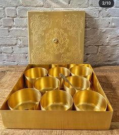 gold colored bowls and spoons in a box on a wooden table next to a brick wall