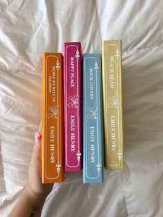 three different colored books sitting on top of a white bed next to a person's hand