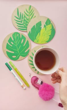 a person holding a cup of coffee next to some green leaf coasters on a pink surface