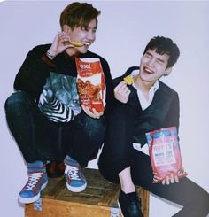 two young men sitting next to each other while eating chips and cereals from bags