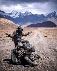 a man riding on the back of a motorcycle down a dirt road with mountains in the background