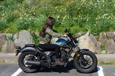 a woman riding on the back of a motorcycle down a street next to grass and rocks