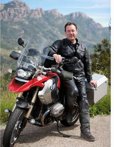 a man sitting on top of a red and silver motorcycle with mountains in the background