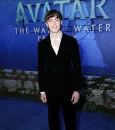 a young man in a black suit standing on a blue carpet