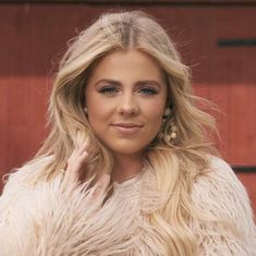 a woman with long blonde hair wearing a fur coat and pearl earrings standing in front of a red wall