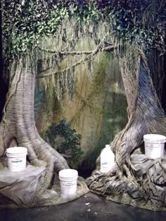 two buckets sitting on top of a rock next to a large tree with moss growing over it