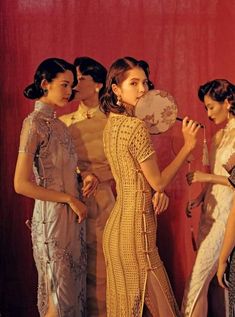 three women standing in front of a red wall and one is holding a fan while the other looks at her reflection