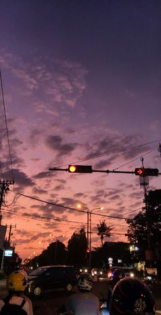 a traffic light hanging over a street filled with cars and motorcycles at dusk, while the sun is setting