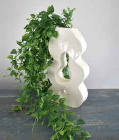 a white vase sitting on top of a wooden table next to a leafy plant