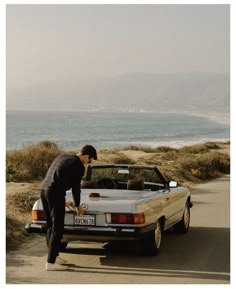 a man standing next to a parked car on the side of a road near the ocean