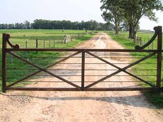 an open gate leading to a dirt road in the middle of a field with grass and trees