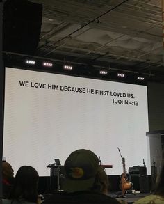 a group of people sitting in front of a large screen with the words we love him because he first loved us