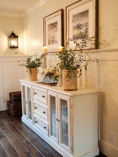 two framed pictures on the wall next to a white cabinet with drawers and vases filled with flowers