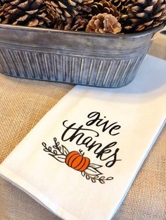 pine cones are sitting in a metal pan on top of a white table cloth that says give thanks