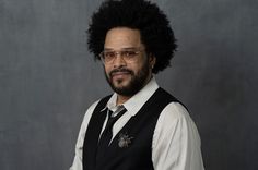 a man with an afro wearing glasses and a black vest is standing in front of a gray background