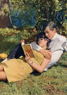 an older woman laying on the grass reading a book to a young boy who is sitting next to her