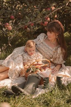 a woman and child sitting in the grass under an apple tree