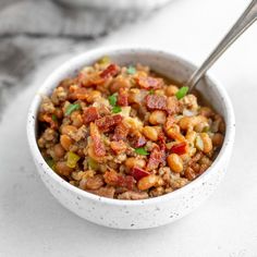 a white bowl filled with beans and meat on top of a marble counter next to a silver spoon