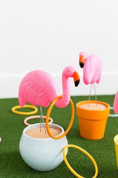 three pink flamingos standing on top of a green grass covered field next to potted plants