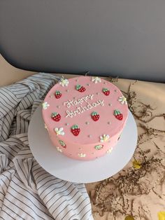 a pink birthday cake sitting on top of a white plate next to a striped blanket