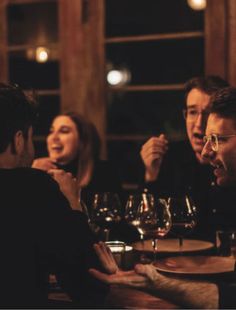 three people sitting at a table with wine glasses in front of them, talking and laughing