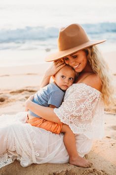 a woman in a hat holding a baby on the beach while wearing a white dress