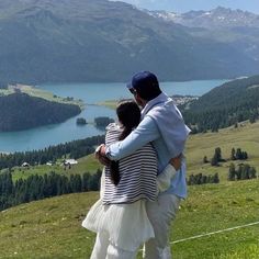 a man and woman standing on top of a lush green hillside next to a lake