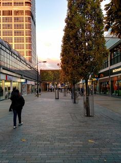 a person is walking down the street in front of some tall buildings and trees with leaves on them