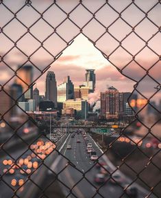 the city skyline is seen through a chain link fence with cars driving on the road