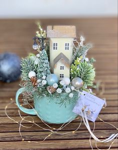 a small house in a teacup filled with greenery and christmas decorations on a wooden table