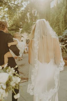 a bride and groom walking down the street with their baby in her arms as they hold each other's hands