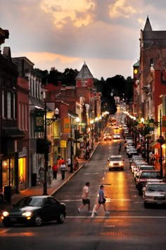 a city street with cars and people walking on the sidewalk at night time, as the sun sets