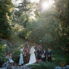 a group of people standing around each other in front of some trees and bushes with flowers